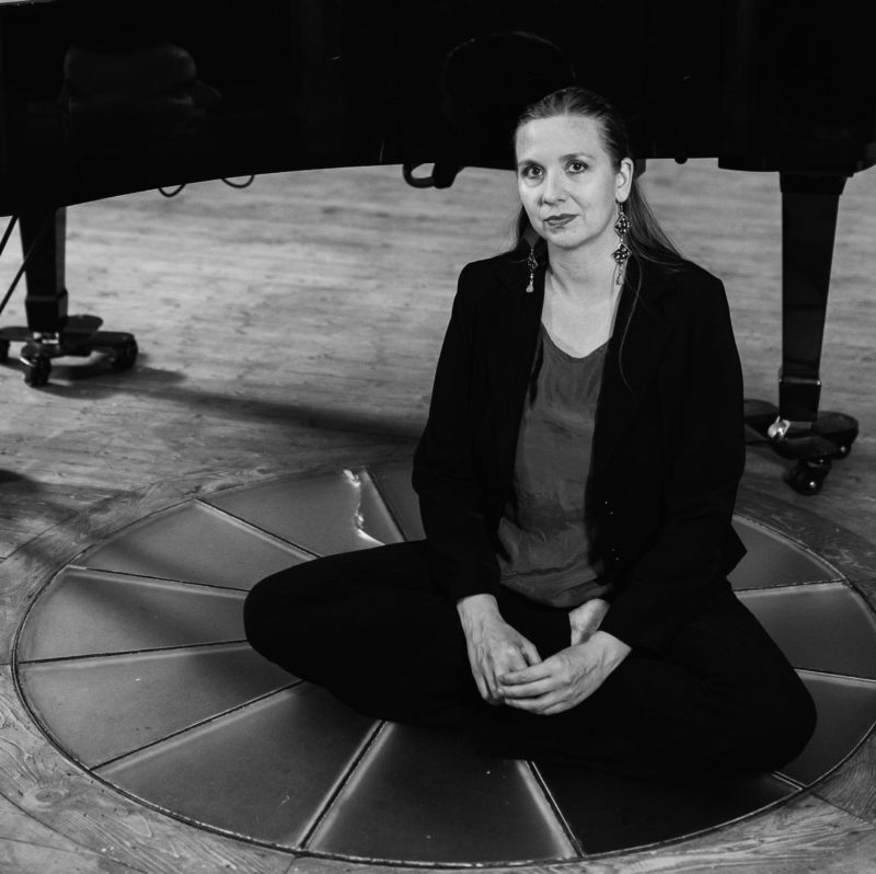 Photo en noir et blanc de la pianiste Viva Sanchez assise à même le sol devant un piano à queue.,